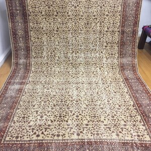 Puede incluir: Una alfombra vintage de color crema con un patrón floral en tonos marrones y rojos. La alfombra tiene un borde con un patrón repetitivo en tonos rojos, marrones y morados.