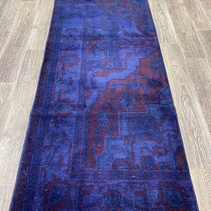 Pu&ograve; includere: Un tappeto runner vintage blu e bordeaux con un design persiano tradizionale. Il tappeto ha un aspetto sbiadito e usurato ed &egrave; fatto di lana.