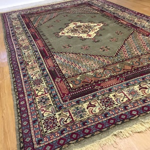 May include: A vintage rug with a green center and a multi-colored border. The rug has a diamond pattern in the center and a floral design in the border. The rug is fringed on all sides.