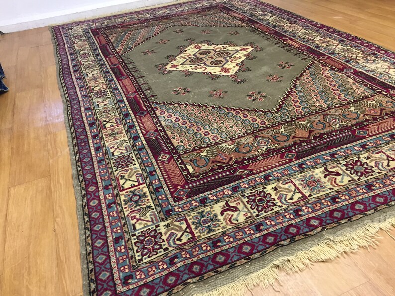 May include: A vintage rug with a green center and a multi-colored border. The rug has a diamond pattern in the center and a floral design in the border. The rug is fringed on all sides.