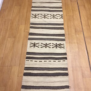 May include: A long, narrow, white rug with brown and black geometric patterns. The rug has a fringe on the ends.