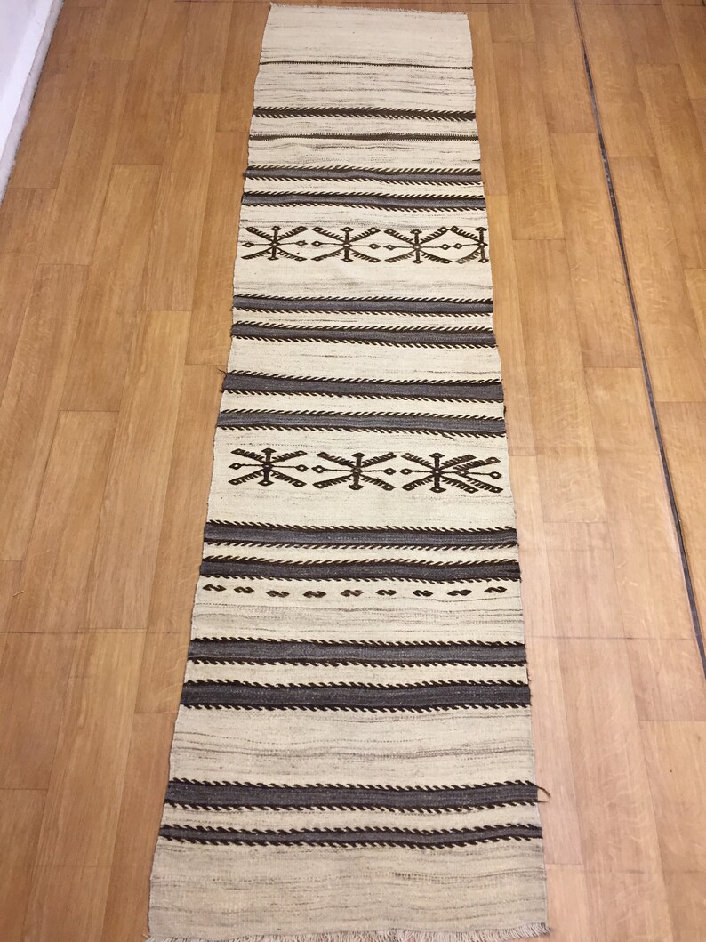 May include: A long, narrow, white rug with brown and black geometric patterns. The rug has a fringe on the ends.