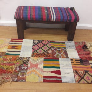 以下が含まれることがあります： 赤、オレンジ、黄色、緑、青、白の様々な幾何学模様が施されたカラフルなパッチワークの敷物。ストライプの布地のクッションが付いた木製のベンチが敷物の前に置かれている。