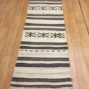 May include: A long, narrow, white and brown woven rug with a geometric pattern. The rug has a fringe on the ends.