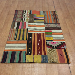 以下が含まれることがあります： さまざまな幾何学模様がカラフルに組み合わされた長方形のパッチワークラグ。オレンジ、茶色、緑、ピンクの色合いで、ストライプ、シェブロン、その他のデザインが混在しています。木製の床に敷かれたラグです。