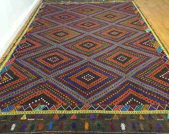 May include: A colorful rug with a geometric diamond pattern in shades of red, orange, blue, green, and brown. The rug has a border with a repeating pattern of triangles and small geometric shapes.