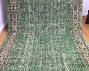 May include: A large, rectangular green rug with a floral pattern and a cream-colored border. The rug has a distressed, vintage look, with faded areas and a worn texture. The border features intricate floral and geometric designs, adding to its antique aesthetic.