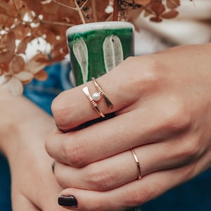 May include: A close-up of a hand wearing two gold rings. The ring on the index finger has a small pearl accent. The ring on the ring finger is a simple band. The hand is holding a green ceramic vase with dried flowers.