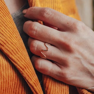 May include: A gold-toned ring with a wavy design. The ring is worn on the index finger of a person's hand.