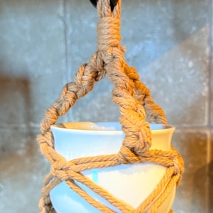 Puede incluir: Un macetero colgante de macramé con una maceta de cerámica blanca y una base de madera. El macramé está hecho de cuerda de color beige y está suspendido de un anillo de metal negro. La maceta está vacía.