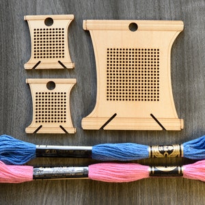 May include: Three wooden embroidery hoops with a grid pattern, two small and one large, and two spools of embroidery floss, one pink and one blue.