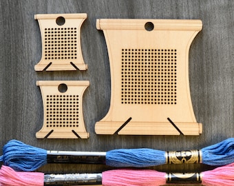 May include: Three wooden embroidery hoops with a grid pattern, two small and one large, and two spools of embroidery floss, one pink and one blue.