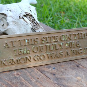 May include: A rectangular bronze plaque with the inscription "AT THIS SITE ON THE 15th OF JULY 2016, POKÉMON GO WAS A BIG DEAL." The plaque is set on a wooden surface, with a skull in the background.