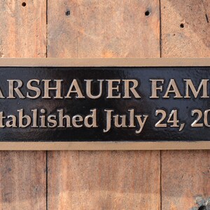May include: A rectangular, bronze-colored plaque with the words "WARSHAUER FAMILY" and "Established July 24, 2009" in raised lettering. The plaque has a dark background and is mounted on a wooden surface.