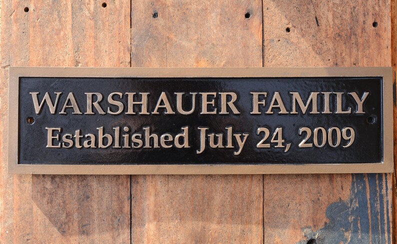 May include: A rectangular, bronze-colored plaque with the words "WARSHAUER FAMILY" and "Established July 24, 2009" in raised lettering. The plaque has a dark background and is mounted on a wooden surface.
