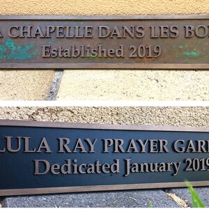 May include: Two rectangular metal plaques. The top plaque reads "LA CHAPELLE DANS LES BOIS Established 2019". The bottom plaque reads "LULA RAY PRAYER GARDEN Dedicated January 2019". Both plaques have bronze lettering.