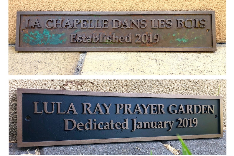 May include: Two rectangular metal plaques. The top plaque reads "LA CHAPELLE DANS LES BOIS Established 2019". The bottom plaque reads "LULA RAY PRAYER GARDEN Dedicated January 2019". Both plaques have bronze lettering.