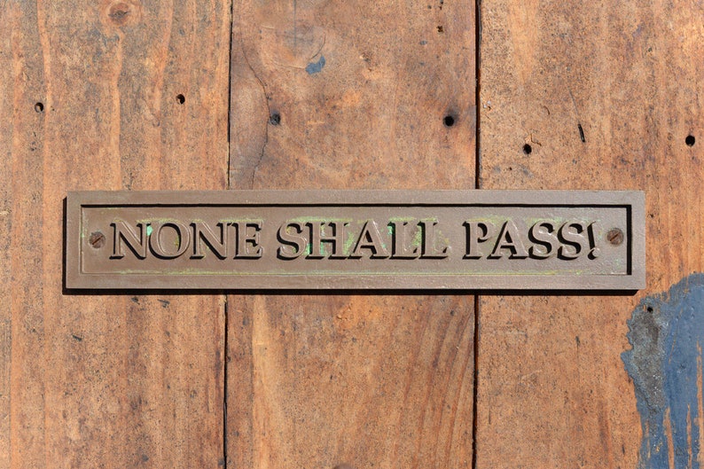 May include: A rectangular bronze plaque with the words "NONE SHALL PASS!" in bold, raised lettering is mounted on a weathered wooden door. The plaque is secured with small screws, and the wood has a natural, aged appearance.