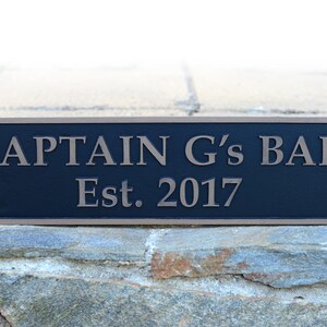 May include: Rectangular black sign with the text "CAPTAIN G's BAR Est. 2017" in a bronze color. The sign has a bronze border and is mounted on a stone wall. The sign is a decorative item.