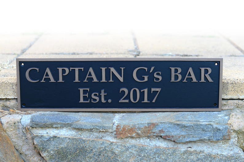 May include: Rectangular black sign with the text "CAPTAIN G's BAR Est. 2017" in a bronze color. The sign has a bronze border and is mounted on a stone wall. The sign is a decorative item.