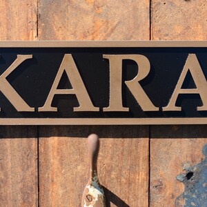 May include: A rectangular sign with the name "KARA" in gold lettering against a black background. The sign has a brown border and is mounted on a wooden surface. A handle is visible below the sign.