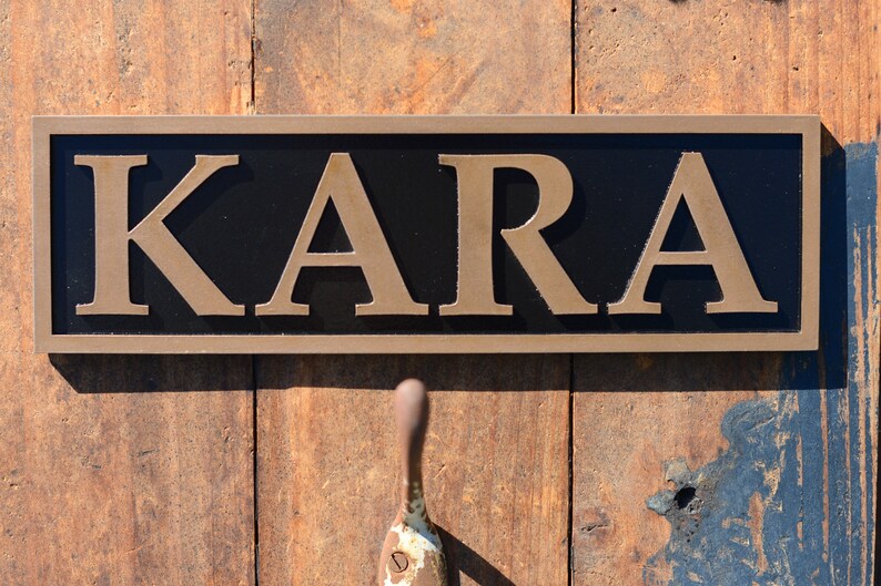 May include: A rectangular sign with the name "KARA" in gold lettering against a black background. The sign has a brown border and is mounted on a wooden surface. A handle is visible below the sign.