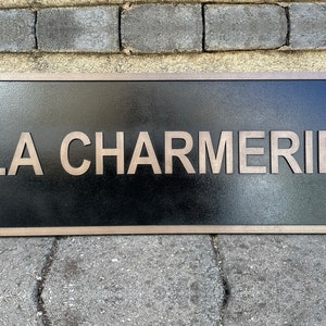 May include: Rectangular black sign with the text "LA CHARMERIE" in a light brown, sans-serif font. The sign has a light brown border and is mounted on a brick surface.