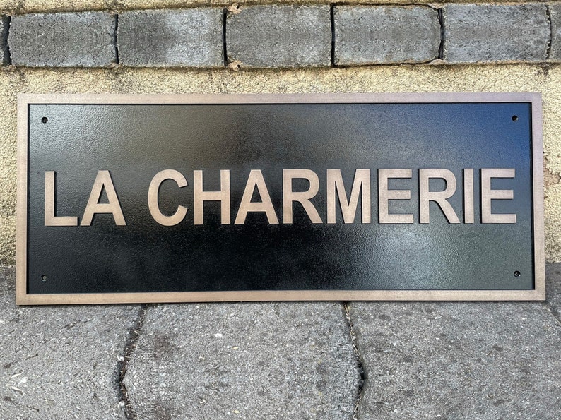 May include: Rectangular black sign with the text "LA CHARMERIE" in a light brown, sans-serif font. The sign has a light brown border and is mounted on a brick surface.