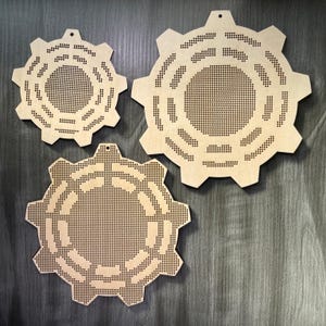May include: Three wooden gear-shaped wall decorations. Each gear has a series of small holes and a larger central opening. The gears vary in size and are made of light-coloured wood, with a natural finish. They are designed to be hung on a wall.