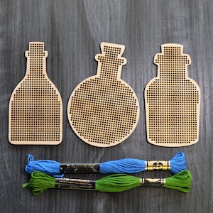 Puede incluir: Tres aros de bordado de madera con forma de botellas. Los aros son de madera marrón claro y tienen un patrón de cuadrícula. Dos de los aros tienen forma de botellas tradicionales, mientras que el tercero tiene forma de botella redonda con tapón. Se incluyen dos ovillos de hilo de bordar, uno azul y uno verde.