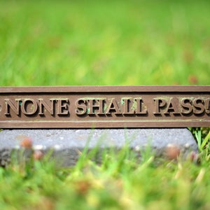 May include: A rectangular bronze plaque with the words "NONE SHALL PASS!" in raised lettering. The plaque is set on a gray stone, with a blurred green grass background.
