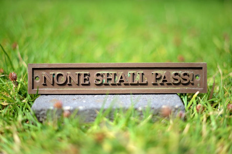 May include: A rectangular bronze plaque with the words "NONE SHALL PASS!" in raised lettering. The plaque is set on a gray stone, with a blurred green grass background.