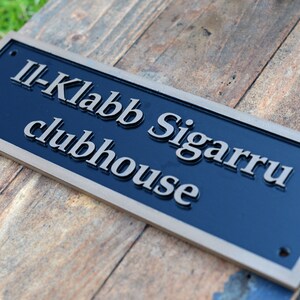 May include: A rectangular sign with the text "II-Klabb Sigarru clubhouse" in silver lettering on a navy blue background. The sign has a gold-colored border and is mounted on a wooden surface.