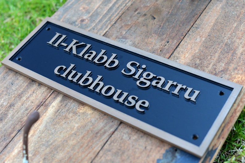 May include: A rectangular sign with the text "II-Klabb Sigarru clubhouse" in silver lettering on a navy blue background. The sign has a gold-colored border and is mounted on a wooden surface.