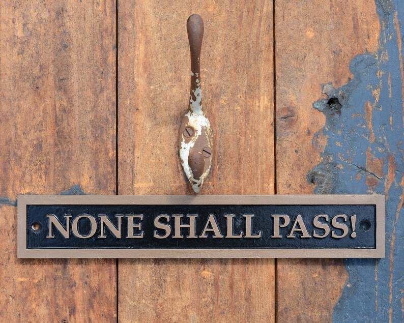 May include: A weathered, brown wooden door with a rusty metal spoon-shaped handle. Below the handle is a rectangular sign with the words "NONE SHALL PASS!" in bronze lettering.