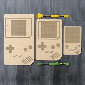 Peut inclure: Trois motifs de consoles de jeux en bois pour broderie, avec des trous pré-percés. Les motifs sont en forme de Game Boy, avec un écran, des boutons et un pavé directionnel. Les motifs sont accompagnés de deux bobines de fil à broder, une jaune et une verte.