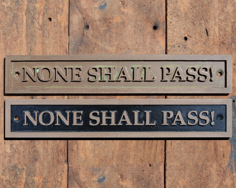 May include: Two rectangular metal signs with the text "NONE SHALL PASS!" are displayed. The top sign is bronze-colored with green oxidation, while the bottom sign is black with bronze lettering. Both signs have mounting holes.
