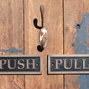 May include: Wooden door with a rusty metal hook and two rectangular signs. The signs read "PUSH" and "PULL" in bronze lettering. The door has a natural wood grain with areas of blue paint.
