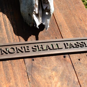 May include: A weathered wooden sign with the words "NONE SHALL PASS!" in raised lettering. The sign is bronze-colored and rests on a rustic wooden surface, with a partial animal skull in the background.