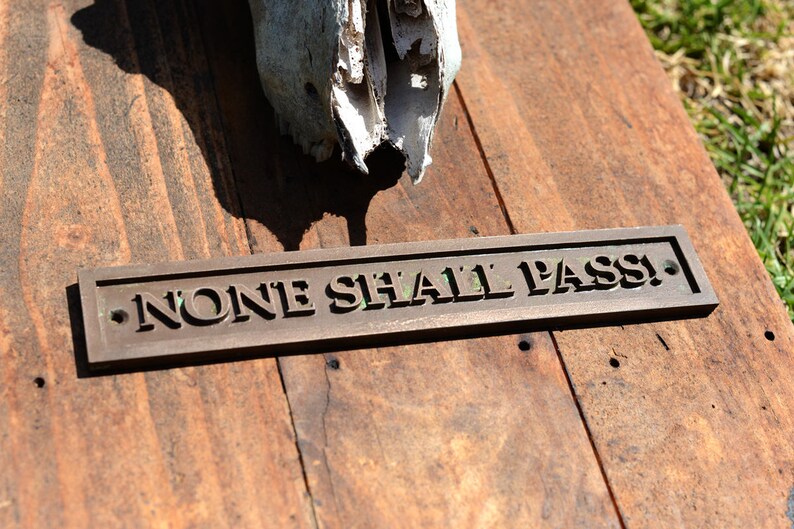 May include: A weathered wooden sign with the words "NONE SHALL PASS!" in raised lettering. The sign is bronze-colored and rests on a rustic wooden surface, with a partial animal skull in the background.