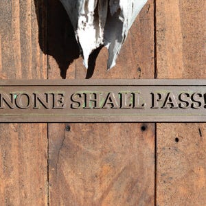May include: A rectangular bronze sign with the words "NONE SHALL PASS!" is mounted on a weathered wooden door. The sign has a slightly aged appearance, with visible screw heads. The wood has a natural, rustic look.