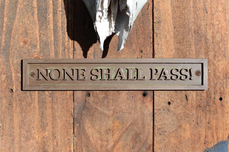 May include: A rectangular bronze sign with the words "NONE SHALL PASS!" is mounted on a weathered wooden door. The sign has a slightly aged appearance, with visible screw heads. The wood has a natural, rustic look.