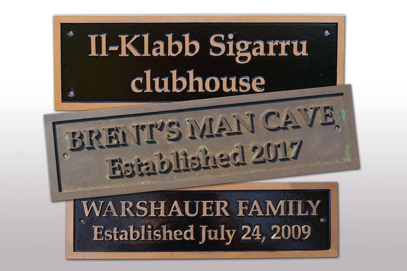 May include: Three rectangular plaques with text. The top plaque reads "Il-Klabb Sigarru clubhouse" in bronze lettering on a black background. The middle plaque says "BRENT'S MAN CAVE Established 2017". The bottom plaque reads "WARSHAUER FAMILY Established July 24, 2009".