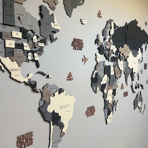 May include: A multi-layered wooden world map wall art piece featuring shades of gray, white, and natural wood tones. The map includes labeled continents, countries, and US states, accented with small wooden icons of airplanes and sailboats.