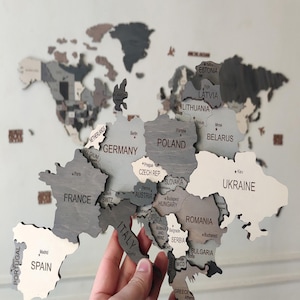 Close-up of dark grey 3D wooden world map — hand holding laser-cut Europe piece showing Germany, France, Poland, Ukraine and surrounding countries