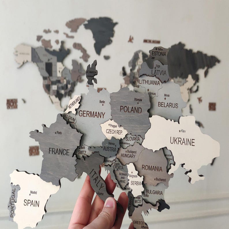 Close-up of dark grey 3D wooden world map — hand holding laser-cut Europe piece showing Germany, France, Poland, Ukraine and surrounding countries