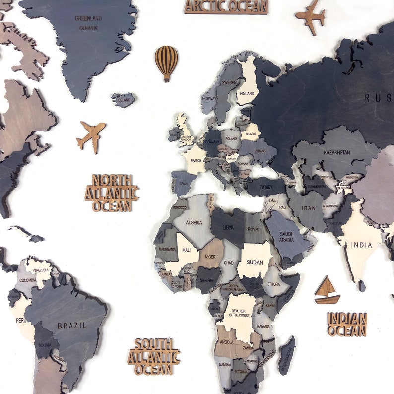 A wooden world map with countries in shades of gray, brown, and beige. The map includes wooden cutouts of an airplane, hot air balloon, and sailboat. The oceans are labeled with wooden text cutouts.