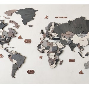 May include: A wooden world map wall decor in shades of gray, brown, and white. The map features individual country cutouts and includes decorative elements like airplanes and boats. The map is mounted on a white wall.