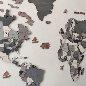 May include: A wooden world map wall decor in shades of gray, beige, and brown. The map is segmented into countries and states, with small wooden sailboats and airplanes scattered around. The words "North Atlantic Ocean" and "Arctic Ocean" are also present.