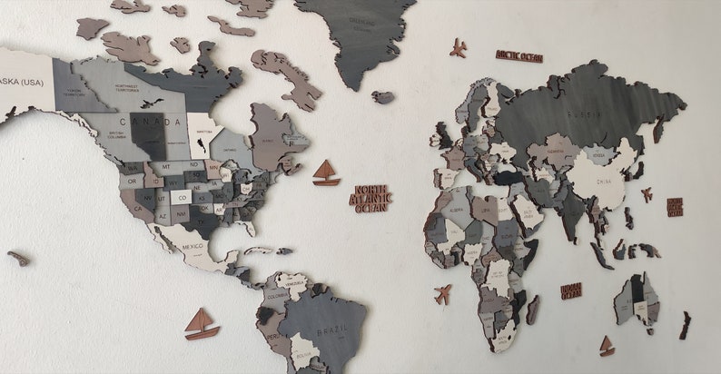 May include: A wooden world map wall decor in shades of gray, beige, and brown. The map is segmented into countries and states, with small wooden sailboats and airplanes scattered around. The words "North Atlantic Ocean" and "Arctic Ocean" are also present.